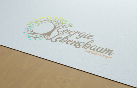 Bild: Ein 3D-Rendering des Logos "Energie Lebensbaum" als Druck auf gestrichenem weißen Papier. Das Logo selbst besteht aus einem Baum mit vollem Blätterdach und weitreichenden Wurzeln die langsam neben dem Stamm nach links zusammenfinden und mit ihm einen Kreis bilden. Nach rechts wächst der Baum in einen handschriftlich anmutenden Schriftzug der "Energie Lebensbaum" liest und bei dem jeder Buchstabe vollständig mit dem nächsten verbunden ist. Rechts abschließend darunter befindet sich der Name "Nathalie Langer" in fester klarer serifenloser Schrift.