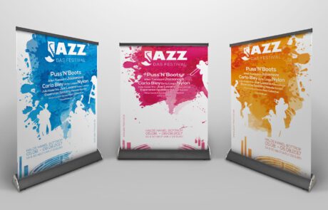 Bild: 3D-Rendering dreier Rollups die mit dem Posterdesign des Jazz Festivals in drei Varianten bedruckt wurden. Darauf befinden sich das Festivaldatum, Lineup und die üblichen Designelemente in Form von Farbsplashes und daraus ausgeschnittenen Silhouetten verschiedener Bandmitglieder. Als Ort ist die Halde Haniel verzeichnet, die sich im unteren Rand des Design je grafisch angedeutet befindet.