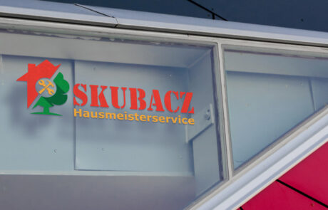 Bild: Das Logo zu Scubacz Hausmeisterservice als Fensterbeschriftung. Es besteht aus einer Bildmarke an der linken Seite, die ein rotes Haus hinter einem grünen Baum mittels eines diagonalen Schnittes verbindet, mit einem runden Freiraum in der Mitte in dem gelb gekreuzt ein Hammer und ein Schraubenschlüssel eingelassen sind. Rechts von der Bildmarke befindet sich der Name "Scubacz" in großer roter Druckschrift, die Aufschrift "Hausmeisterservice" befindet sich in Gelb in gleicher Länge darunter.